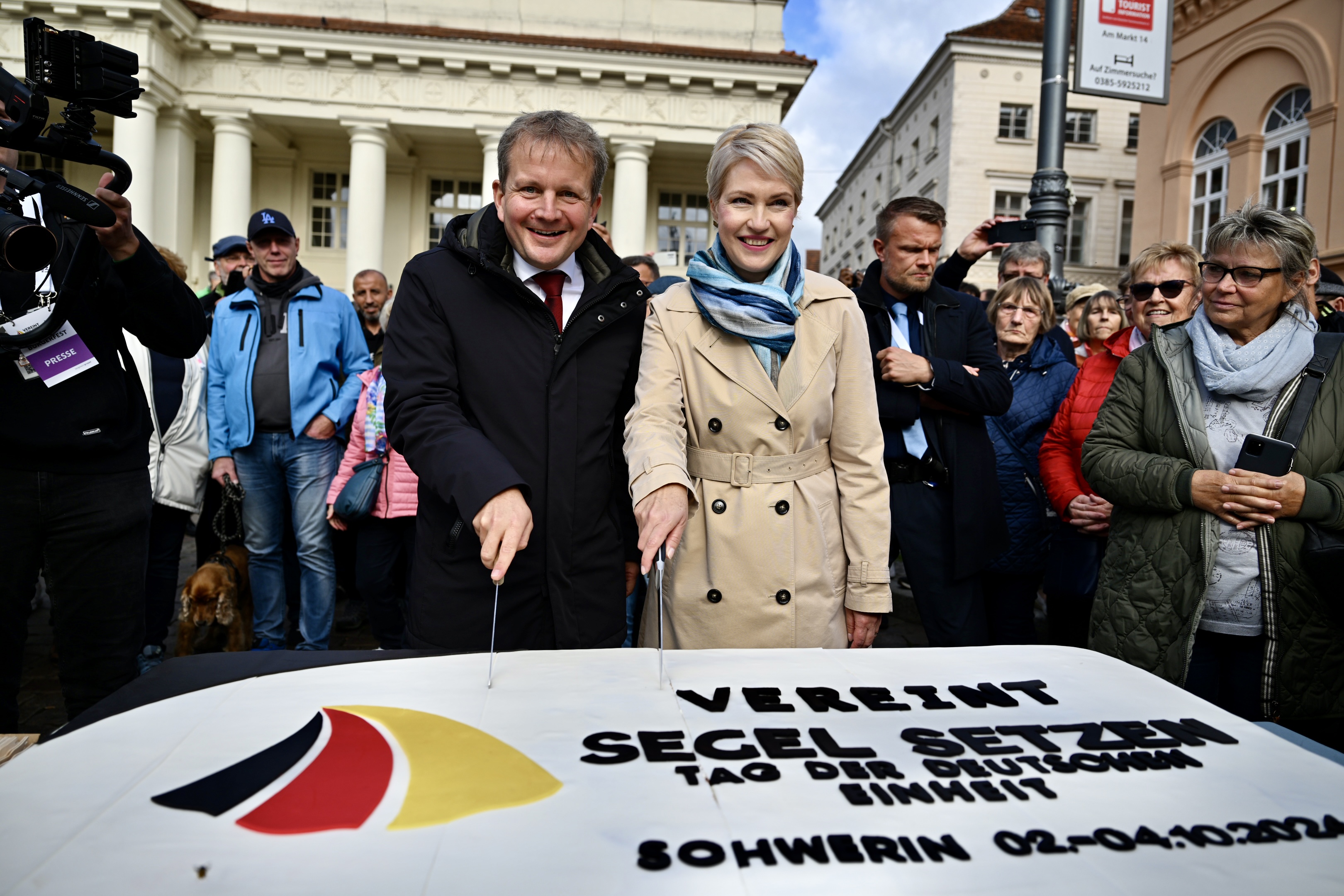 Das Bild zeigt zwei Personen, einen Mann und eine Frau, die gemeinsam eine große Torte anschneiden. Die Torte ist mit einer deutschen Flagge und dem Schriftzug "VEREINT SEGEL SETZEN – Tag der Deutschen Einheit – Schwerin 02.-04.10.2023" verziert. Im Hintergrund sind mehrere Menschen zu sehen, die an einer Veranstaltung teilnehmen. Die Personen lächeln und tragen wetterangepasste Kleidung – der Mann eine dunkle Jacke, die Frau einen beigefarbenen Mantel mit einem blauen Schal. Die Umgebung deutet auf einen ö