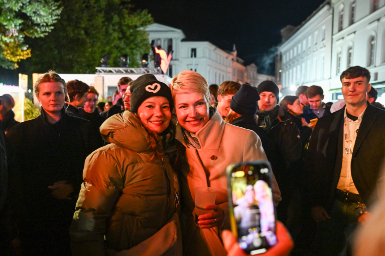 Manuela Schwesig umarmt eine rau in einem dicken Annorack. Beide lächeln in eine Handykamera. Im Hintergrund sind viele Menschen.