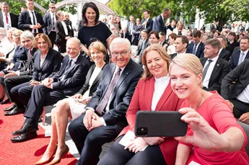 Festlich gekleidete Menschen sitzen in Reihen. Im rechten Vordergrund hält Manuela Scheswig ein Handy für ein Selfie in die Höhe.