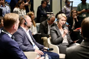 Manuela Schwesig trägt Kopfhörer und spricht in ein Mikrofon. Sie sitz in einem Sessel umgeben von mehreren Menschen in einem Studio.