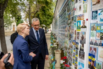 Manuela Schwesig steht mit einem Mann vor einer Wand mit Porträts von Verstorbenen, die am Boden von Blumen gesäumt ist.