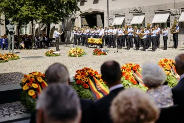 Eine offizielle Gedenkzeremonie mit mehreren Menschen auf einem großen Platz. Blumen liegen nieder. Ein Orchester spielt Musik.