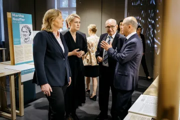 Manuela Schwesig steht mit mehreren Menschen darunter Olav Scholz in einem Ausstellungsraum umgeben von Informationstafeln.