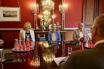 Schwesig sitzt an einem breiten Konferenztisch in einem königlichen Raum. Links neben ihr sitzt eine blonde Frau, links neben ihr ein älterer Herr.