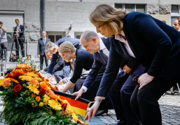 Mehrere Personen in Anzügen legen Kränze auf einem Platz nieder.