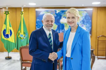 Ein älterer Herr in dunkelblauem Anzug schüttelt Manuela Schwesig in hellem Anzug die Hand. Beide lächeln in die Kamera. Im Hintergrund ein Saal mit brasilianischen Flaggen.