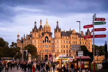 Das Bild zeigt das beleuchtete Schweriner Schloss bei Dämmerung. m Vordergrund sind viele Menschen zu sehen, die sich auf einem öffentlichen Platz vor dem Schloss versammeln. Es gibt verschiedene Imbissstände, die Speisen wie Wurst und Crêpes anbieten. 