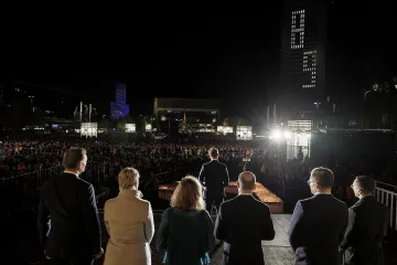 Mehrere Menschen stehen mit dem Rücken zur Kamera auf einer Bühne vor mehreren hundert Menschen. Es ist dunkel.