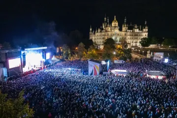 Tausende Menschen stehen vor dem Schweriner Schloss. Auf einer Bühne am linken Bildschirmrand findet ein Programm statt.